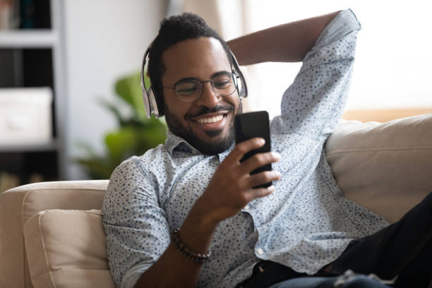 Happy african man wearing headphones listening mobile music on couch Happy african man wearing wireless headphones listening to music or audio book on smartphone relaxing on sofa, smiling young afro american guy enjoying phone player sound lounge on couch at home radio broadcasting photos stock pictures, royalty-free photos & images