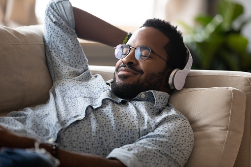 Serene satisfied young adult african american man lying on comfortable sofa wear wireless headphones enjoy listen modern music audio book with eyes closed meditate relax feel no stress chill at home