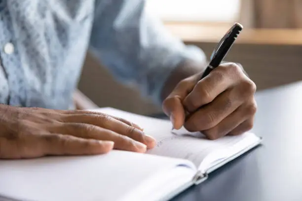 Close up view of african left-handed businessman writing in notebook, american male hands holding pen making notes planning new appointments information in organizer personal paper planner at desk