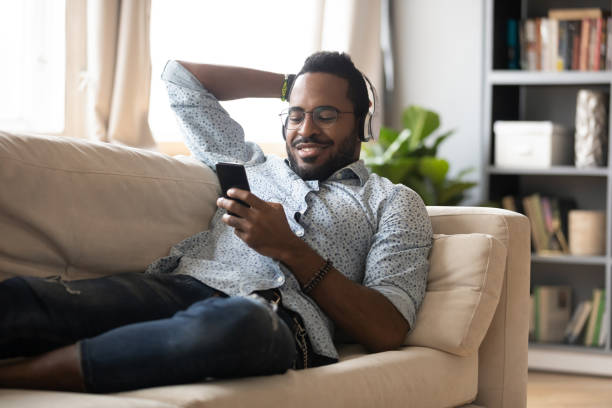 Relaxed african man wear headphones chill on sofa listening audiobook Relaxed millennial african man wear wireless headphones chill lying on sofa listening audiobook on smartphone, smiling young adult afro american guy rest on couch enjoying mobile music in app at home podcast mobile stock pictures, royalty-free photos & images