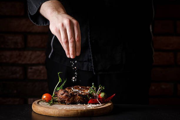 고기를 요리의 개념. 요리사는 검은 색 배경에 요리 스테이크에 소금을 요리, 레스토랑 메뉴에 대한 로고 아래 장소. 음식 배경 이미지, 복사 공간 텍스트 - cut up 이미지 뉴스 사진 이미지