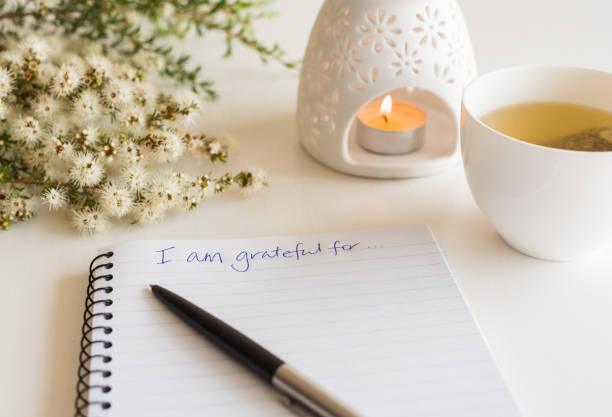 Notebook with "I am grateful for" in handwritten text Close up of handwritten text "I am grateful for..." in foreground with notebook, pen,  cup of tea, flowers and oil burner in soft focus (deliberate angle) low key photos stock pictures, royalty-free photos & images