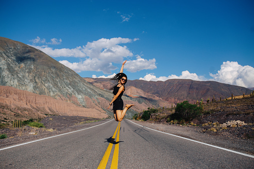 Young woman jumping on an empty road in the middle of nowhere, with dramatic mountains stretching in the background, Salta, Argentina.