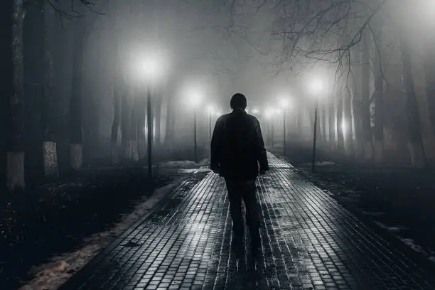 Sad man alone walking along the alley in night foggy park. Back view.