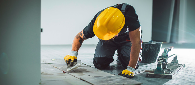 A bearded man in overalls working on a tile floor, filling the joints with grout.