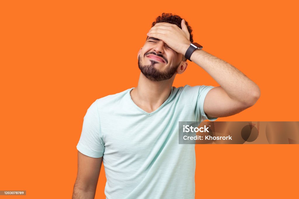 Facepalm. Portrait of desperate brunette man covering face with hand. indoor studio shot isolated on orange background Facepalm. Portrait of desperate brunette man with beard in white t-shirt covering face with hand, feeling sorry and blaming himself for the mistake. indoor studio shot isolated on orange background Men Stock Photo