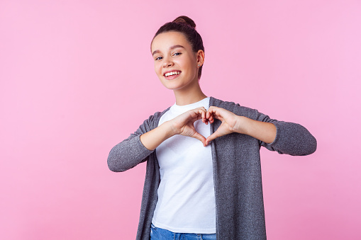 Portrait of lovely brunette teenage girl with bun hairstyle in casual clothes showing heart shape with hands and smiling at camera, romance in adolescence. indoor studio shot isolated, pink background
