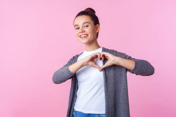 Portrait of lovely brunette teenage girl showing heart shape with hands. indoor studio shot isolated, pink background Portrait of lovely brunette teenage girl with bun hairstyle in casual clothes showing heart shape with hands and smiling at camera, romance in adolescence. indoor studio shot isolated, pink background peace sign gesture photos stock pictures, royalty-free photos & images