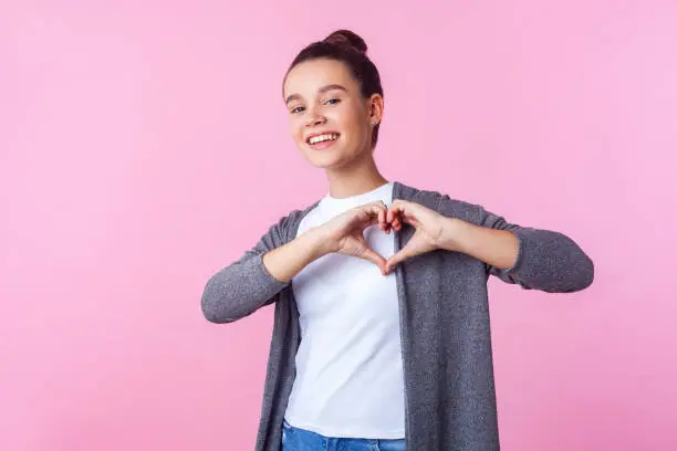 Photo of Portrait of lovely brunette teenage girl showing heart shape with hands. indoor studio shot isolated, pink background