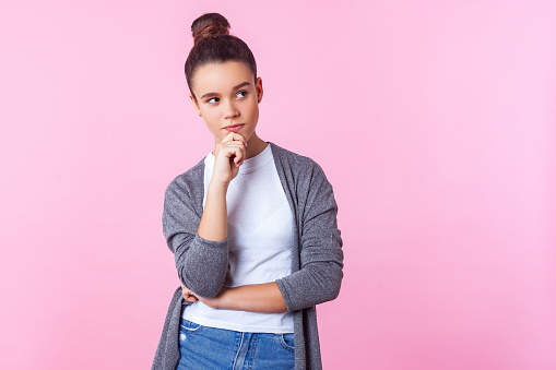 Let me think! Portrait of pensive cute brunette teenage girl with bun hairstyle in casual clothes holding hand on chin and thinking, pondering idea. indoor studio shot isolated on pink background
