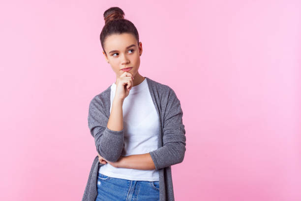 Let me think! Portrait of pensive cute brunette teenage girl holding hand on chin and thinking. isolated on pink background Let me think! Portrait of pensive cute brunette teenage girl with bun hairstyle in casual clothes holding hand on chin and thinking, pondering idea. indoor studio shot isolated on pink background uncertainty photos stock pictures, royalty-free photos & images