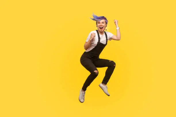 Yes i did it! Portrait of enthusiastic lively hipster girl with violet hair in overalls jumping in air with raised fists, showing yes i did it gesture, life energy. yellow background, studio shot