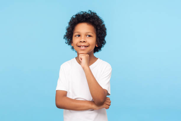 retrato de niño feliz pensativo en camiseta de pie con mirada pensativa y tocando la barbilla, imaginando algo divertido y sonriente - t shirt child white portrait fotografías e imágenes de stock