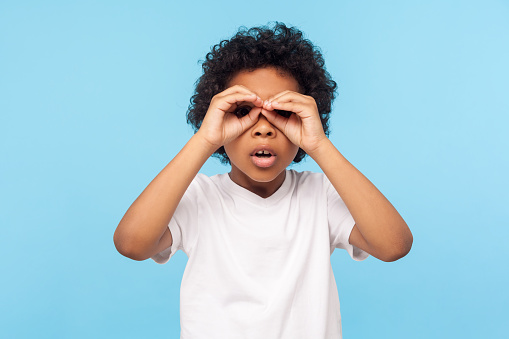 Curious child exploring world. Portrait of inquisitive nosy little curly boy looking through fingers shaped like binoculars and expressing amazement. indoor studio shot isolated on blue background