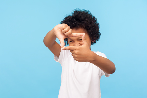 Portrait of curious nosy little boy in T-shirt looking through photo frame shape with fingers, focusing zooming at camera