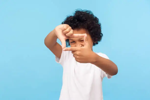 Photo of Portrait of curious nosy little boy in T-shirt looking through photo frame shape with fingers, focusing zooming at camera