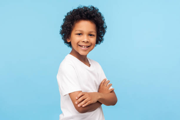 retrato de menino feliz e confiante de camiseta branca de pé com as mãos cruzadas e sorrindo para a câmera - t shirt child white portrait - fotografias e filmes do acervo