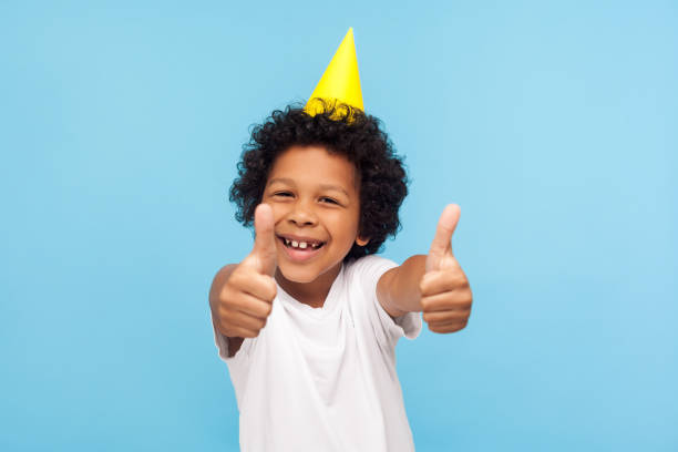 kciuki do przyjęcia urodzinowego! podekscytowany niesamowity radosny mały chłopiec z zabawnym stożkiem na głowie pokazującym jak gest - thumbs up child success winning zdjęcia i obrazy z banku zdjęć