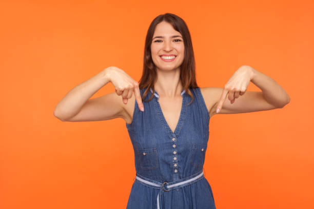 Attention, advertising! Cheerful happy brunette woman smiling and pointing finger down Attention, advertising! Cheerful happy brunette woman in denim dress smiling and pointing finger down, showing below place for promotional text. indoor studio shot isolated on orange background beckoning photos stock pictures, royalty-free photos & images