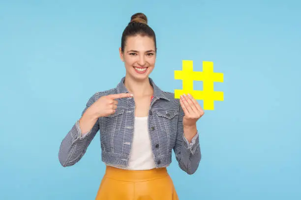 Photo of Optimistic happy attractive woman with hair bun in stylish outfit pointing at big yellow hashtag symbol and smiling at camera