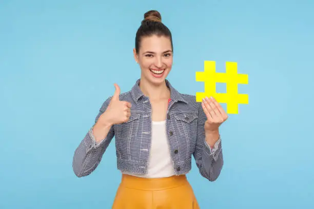 Photo of Like to popular blog and trends. Joyful attractive woman with hair bun in stylish outfit holding big yellow hashtag symbol