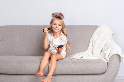 Cute adorable little girl with curlers in hair sitting on sofa and doing make up, smiling at camera, applying face powder with brush, child using cosmetics at home, having fun, beauty and fashion