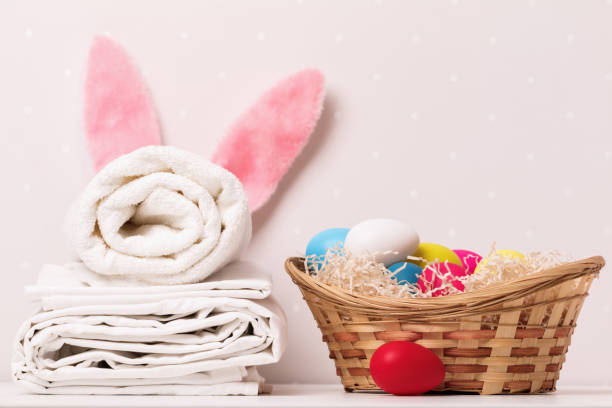 a close-up of a stack of clean white bedding, towels and easter bunny ears, a basket of eggs on a table, against a background of light walls - material data sheets imagens e fotografias de stock