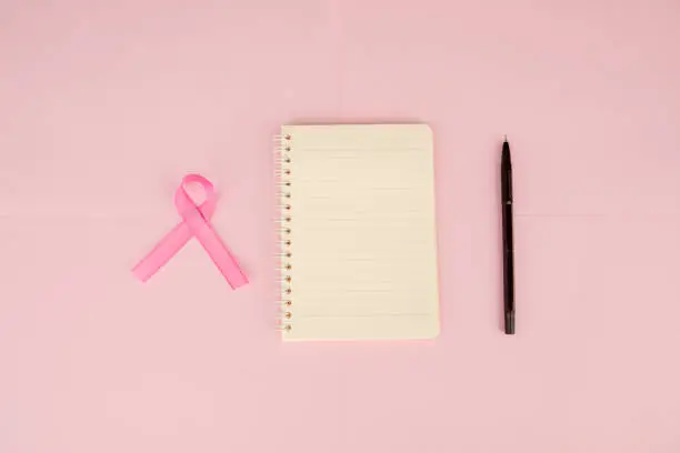 Flat layout of a pink ribbon besides a notebook and a pen, isolated in pink paper background