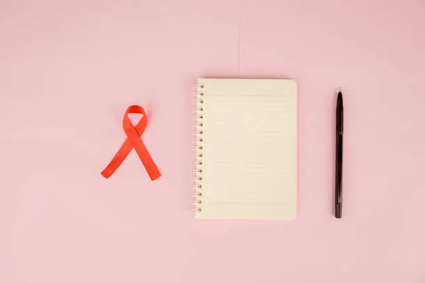 Flat layout of a red ribbon besides a notebook and a pen, isolated in pink paper background