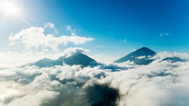 Aerial view of mountain peaks surrounded by cumulus clouds with sun and blue sky background