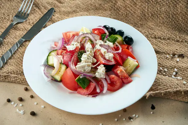 Photo of Classic Greek salad with tomatoes, onions, cucumber, feta cheese and black olives in white plate on a light background. Close up view. Food flat lay