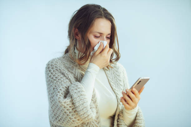 woman wiping nose with napkin ordering pills in online shop ill elegant middle age woman in roll neck sweater and cardigan wiping nose with napkin ordering pills in pharma online shop with smartphone app isolated on winter light blue background. allergy medicine stock pictures, royalty-free photos & images