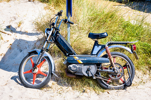 Tregastel, France - August 7, 2019: Vintage Peugeot 103 SP black moped from the early 1980s with red Rigida Leleu 5-spoke wheels leaning against a pole in the sun at the beach.