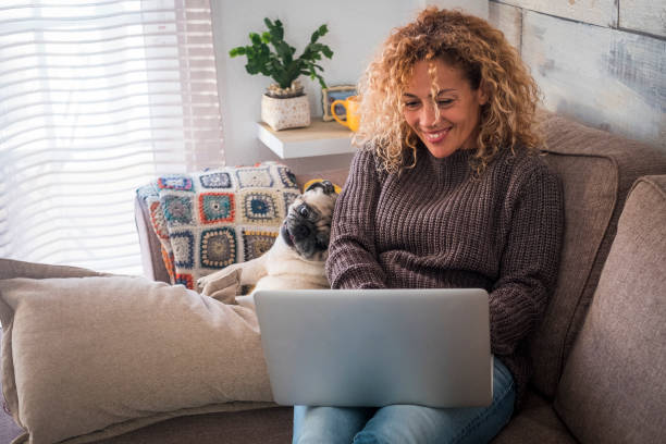 cheerful and nice couple with people and animal - beautiful culry adult woman with funny dog pug at home using personal computer laptop sitting on the sofa together looking the screen - furniture internet adult blond hair imagens e fotografias de stock