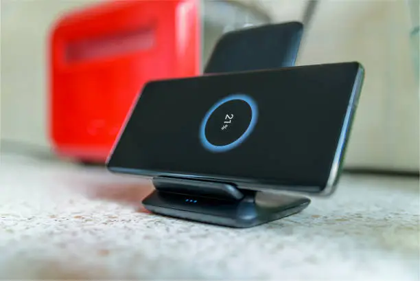 Photo of smartphone wireless charging on charging stand with 21 percent icon on screen on kitchen tabletop next to toaster