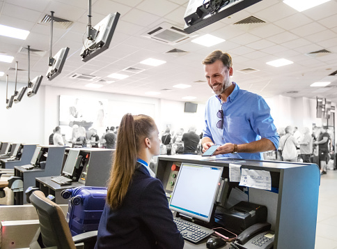 Man standing at check-in counter at the airport and talking with young woman. Defocused crowd in the background.