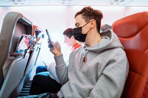 Man sitting on an airplane using his phone while wearing a surgical mask.