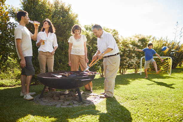 famille appréciant la nourriture grillée, le jeune fils joue au football à l'arrière-plan. - barbecue foot photos et images de collection