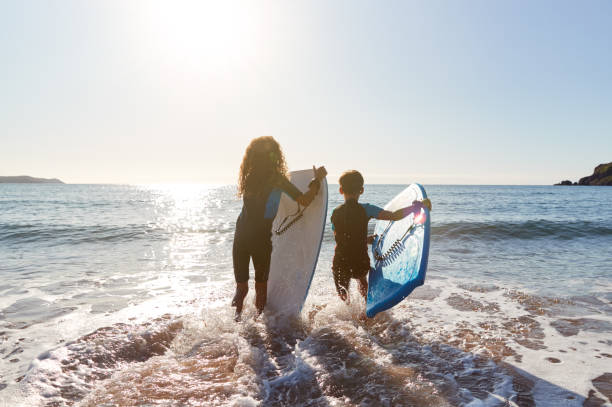 vista trasera de dos niños que llevan trajes de neopreno corriendo hacia el mar con bodyboards on beach vacation - family beach vacations travel fotografías e imágenes de stock