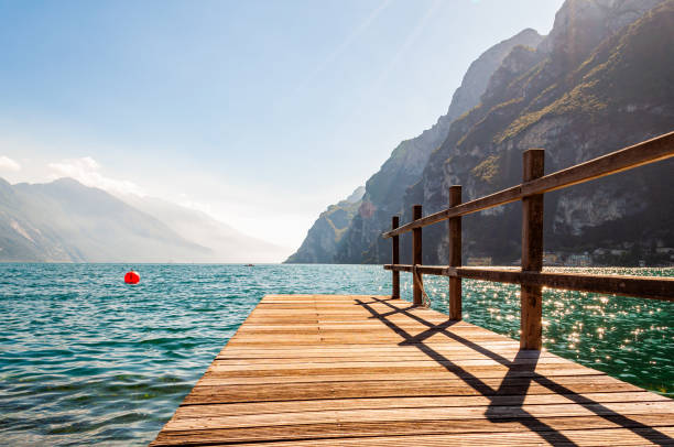 scenic view on wooden planks pier with railings built on northern shore of beautiful garda lake in lombardy, italy surrounded by high dolomite mountains and crystal clear blue water of the lake. riva - buoy horizontal lake sailing imagens e fotografias de stock