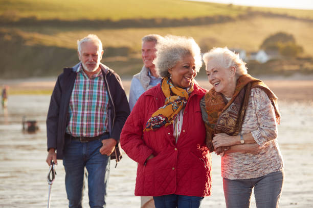 grupo de sonrientes amigos mayores caminando brazo en brazo a lo largo de la costa de winter beach - tercera edad fotografías e imágenes de stock