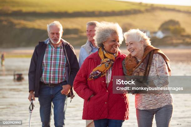 Group Of Smiling Senior Friends Walking Arm In Arm Along Shoreline Of Winter Beach Stock Photo - Download Image Now