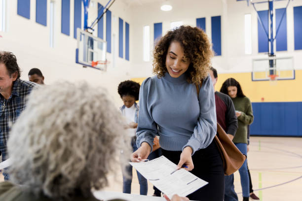 Unrecognizable mature woman assists mid adult woman with voting document The unrecognizable mature adult female polling place volunteer assists the mid adult woman with the document she needs to be aloe to vote.  Other voters use smart phones while waiting in line. democratic party usa photos stock pictures, royalty-free photos & images