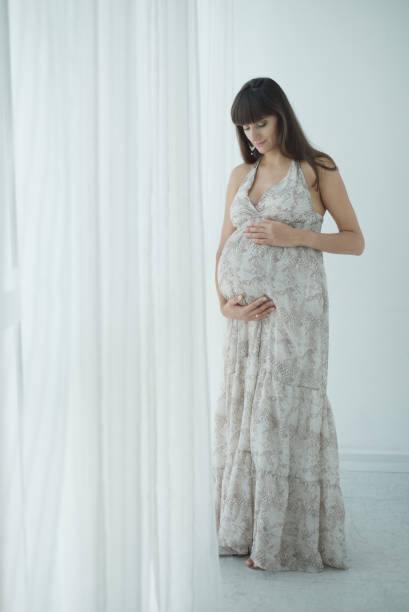 Smiling pregnant woman expecting child standing at the window and caressing her belly Image of happy pregnant woman touching her big belly. Happy pregnant European woman with big tummy is relaxing. Pregnancy, motherhood, people and expectation concept. Complete family. olivia mum stock pictures, royalty-free photos & images