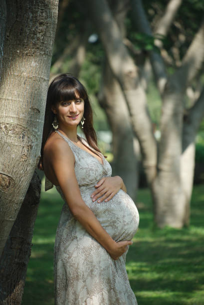 Smiling woman expecting child standing reclining on the tree and caressing her belly Image of happy pregnant woman touching her big belly. Happy pregnant European woman with big tummy is relaxing. Pregnancy, motherhood, people and expectation concept. Complete family. olivia mum stock pictures, royalty-free photos & images