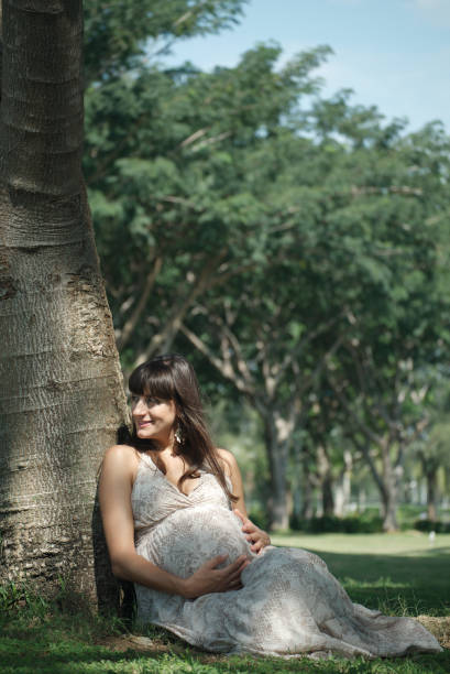 Smiling woman expecting child siting on the lawn near the tree and caressing her belly Image of happy pregnant woman touching her big belly. Happy pregnant European woman with big tummy is relaxing. Pregnancy, motherhood, people and expectation concept. Complete family. olivia mum stock pictures, royalty-free photos & images