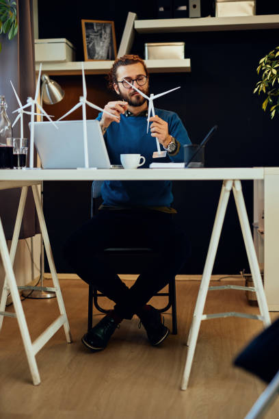 istock Young innovative caucasian bearded engineer sitting at his modern office and holding windmill model. Sustainable concept. 1203202510