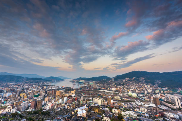 A calm view of Nagasaki Harbor featuring shimmering water, distant mountains, and the silhouette of a working shipyard with cranes. The bright afternoon light reflects across the sea, creating a peaceful coastal atmosphere. Ideal for themes related to Japan travel, port cities, maritime industry, and waterfront scenery.