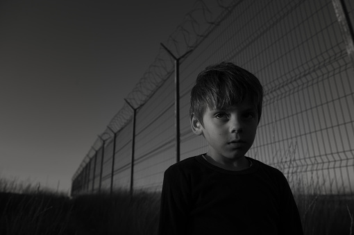 portrait of upset poor little refugee child in black shirt standing near long and high fence with barbed wire on state border on sunset