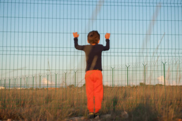 illegal immigration displaced persons camp concept of little poor orphan refugee boy in black and orange clothes holding fence with barbed wire in desert on state border illegal immigration displaced persons camp concept of little boy in black and orange clothes holding fence with barbed wire in desert on state border war zone stock pictures, royalty-free photos & images